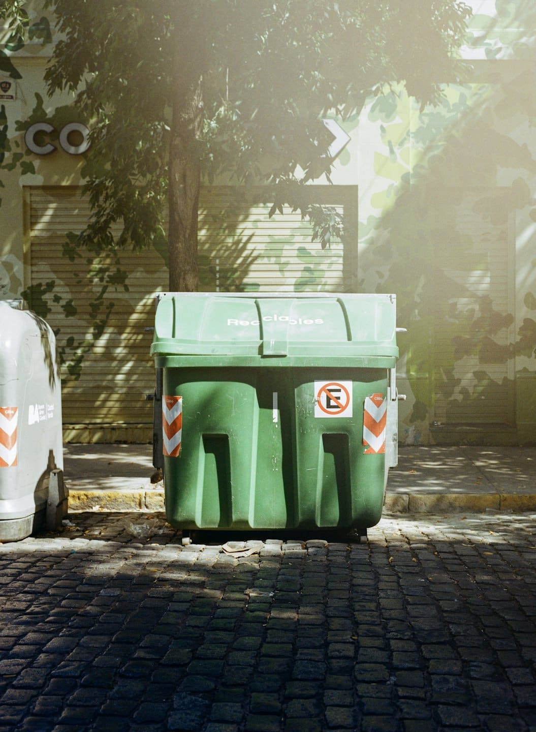 A green recycling bin sits on a cobblestone city street.